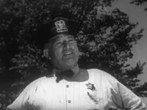 A smiling man in a light shirt and cap stands outside against a tree background.