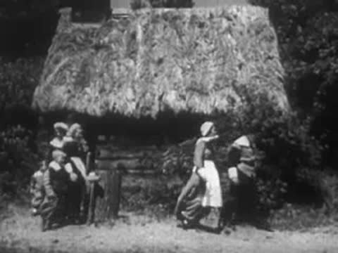 People in colonial-era attire walking near a traditional thatched-roof cottage.