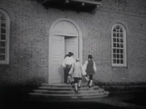 People in historical attire walking up steps toward a large building entrance.