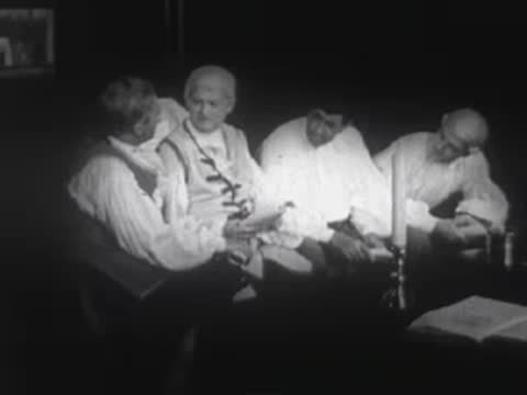 Men in period clothing seated around a table indoors with candles lit.