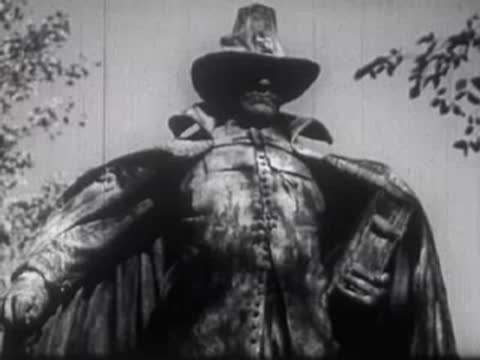 A statue of a historical figure in a cloak and hat outdoors with trees.