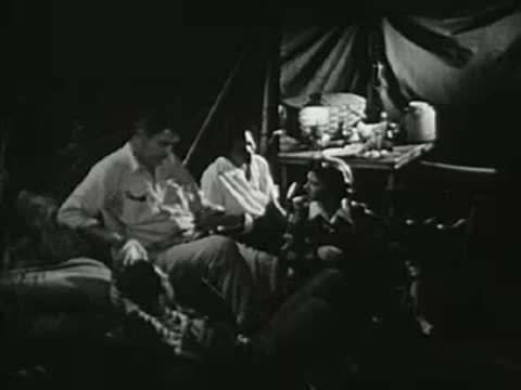 Campsite scene at night with people gathered by a tent and lantern