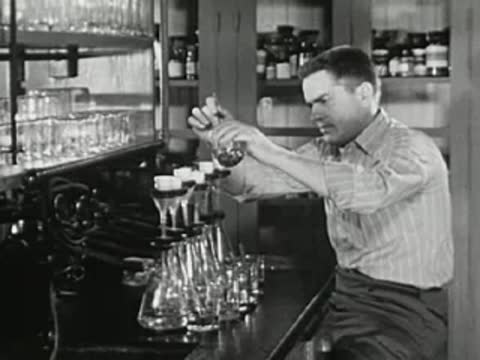 Student swirling liquid in a conical flask, laboratory setting with shelves and glassware