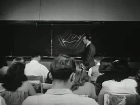 Teacher lecturing in front of a chalkboard with diagram, students seated in lecture hall