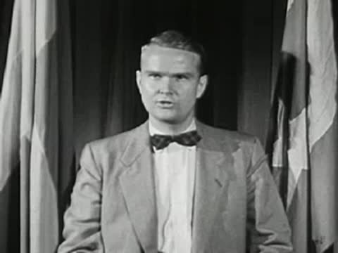 Man in suit and bow tie speaking in front of a curtained backdrop, television broadcast