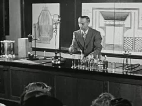 Man adjusting scientific apparatus on a laboratory bench in front of a class