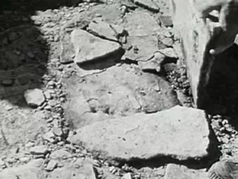 Hands removing stones and debris to reveal a flat rock or paved surface on dirt ground
