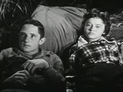 A young man and woman sitting together outdoors at night, looking thoughtfully ahead.