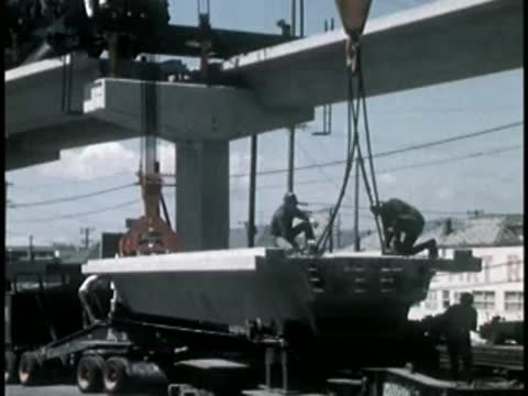 Construction workers guiding a heavy precast concrete beam being lifted by crane.