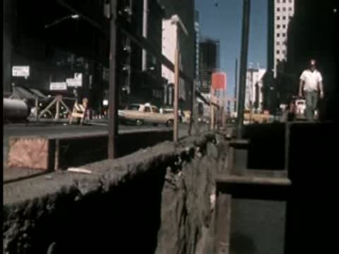 A man standing near a deep excavation trench on an urban street.