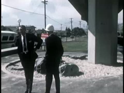 Two men in suits conversing near a highway support pillar outdoors.