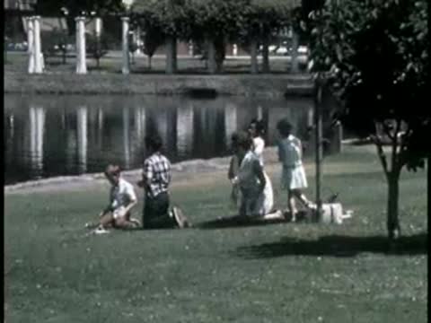 Family group relaxing and playing on a grassy park lawn by water.