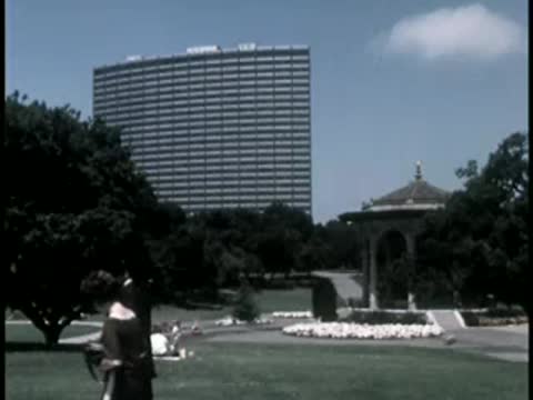 Park scene with tall modern building in background and people sitting on grass.