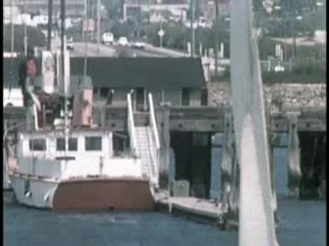 Small sailboat moving on water with buildings and hills in background.