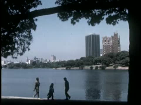 People walking along a path next to a waterfront with city buildings in background.