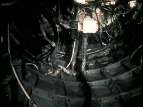 A worker adjusts cables in a dark underground construction tunnel with ribbed lining.