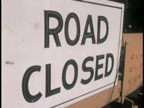 A road closed sign stands near a street construction site with buildings behind.