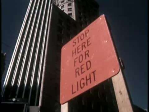 A red traffic sign reading stop here for red light stands against city buildings.