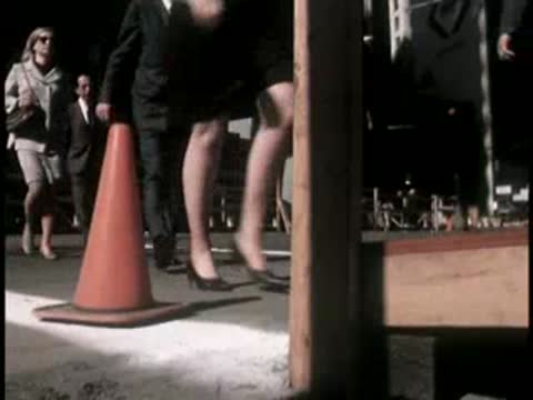 Pedestrians walk past a construction cone and wooden barricade on a city street.