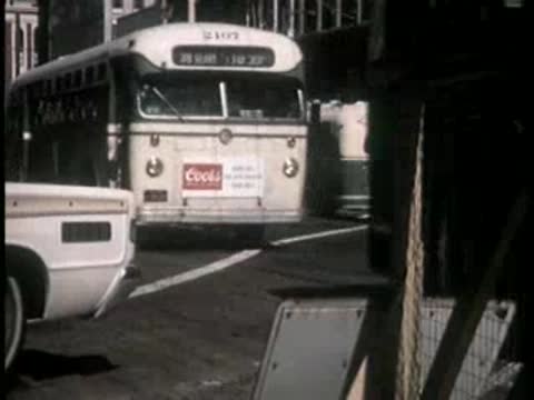 A bus drives down a city street past construction barriers and parked cars.