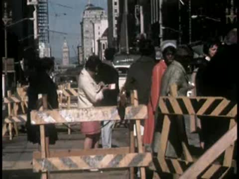 Pedestrians walk past a wooden street barricade with urban buildings in the background.