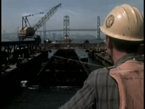 Worker in a hard hat and life jacket watching construction activity from a barge.