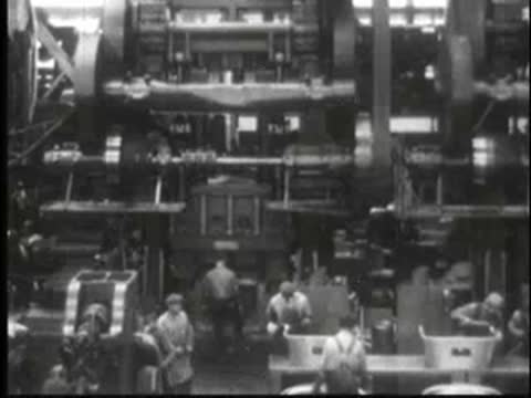 Busy factory floor with complex machinery, overhead belts, and working operators.