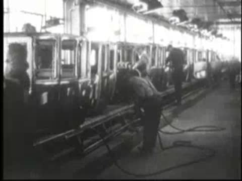Long assembly line with car chassis and workers performing tasks in a factory.