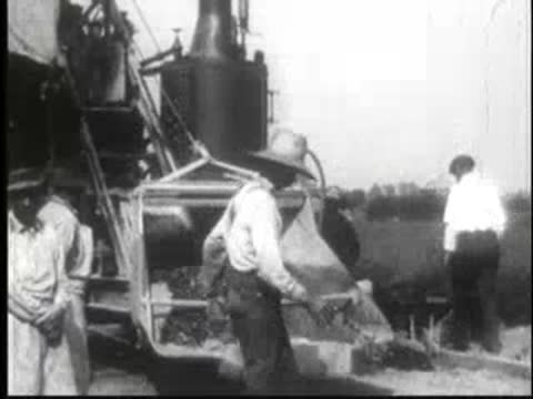 Men operating early road paving machinery dumping gravel on road.