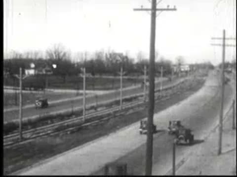 Vehicles driving along a multi-lane rural road intersection with utility poles.