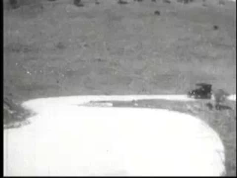 Old car driving along a curved paved road through grassy hills.