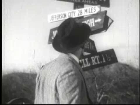 Man in fedora and suit looking at wooden directional signposts.