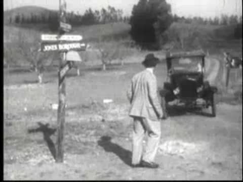 Man in suit walking away from parked vintage car near signposts.