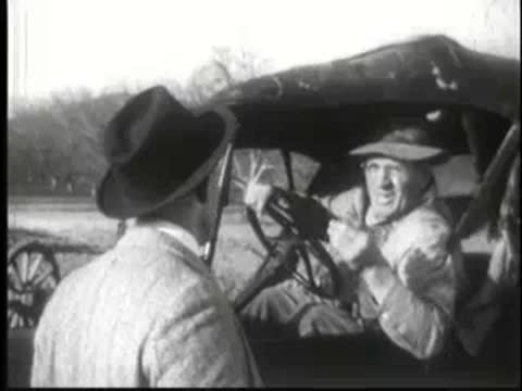 Two men talking inside vintage car, one gesturing with hand.