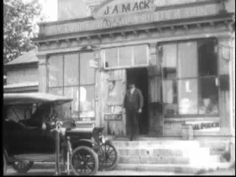 Old car parked outside storefront, J.A. Mack building in background.