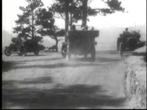 Vintage cars parked near trees with mountains in the background.