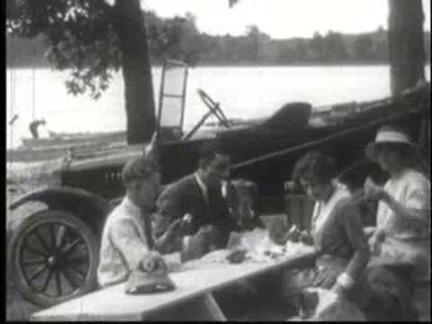 People having a picnic at a table by the water with a car behind them.
