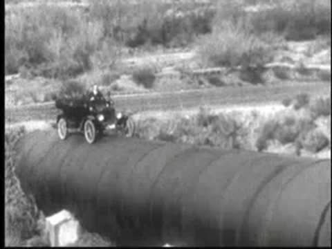 Vintage car driving across the top of a massive cylindrical pipeline.