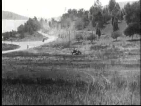 Vintage car driving across a grassy field chased by motorcycles in the distance.