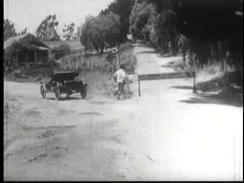 Vintage car crashing through a wooden barrier gate on a rural road.