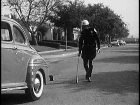 Police officer placing evidence marker on street behind vintage car, 1960s