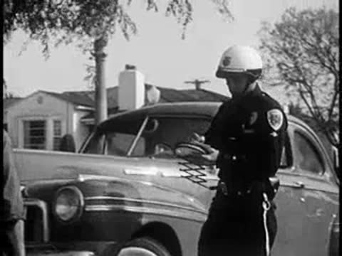 Police officer writing ticket by vintage car on suburban street, 1960s