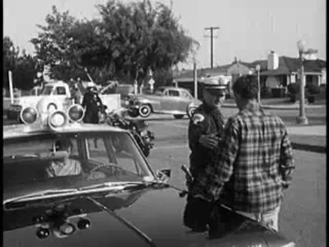 Police officer talking to driver by vintage car on suburban street, 1960s
