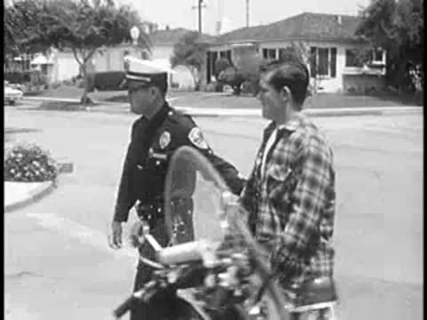 Police officer walking a young man past a parked motorcycle.