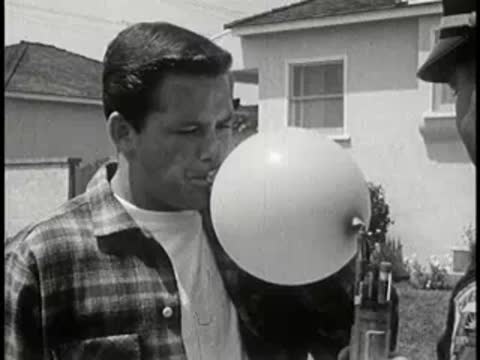 Man blowing up a balloon outdoors wearing a plaid shirt, vintage educational film.