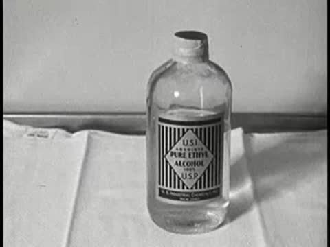 A glass bottle of pure ethyl alcohol on a table with a white cloth, vintage educational film.