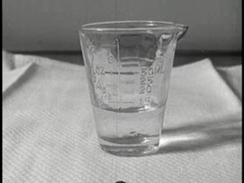 A measuring glass container with liquid on a white cloth on a table, vintage educational film.