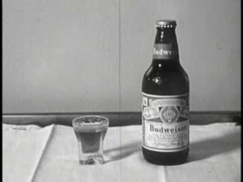 A bottle of Budweiser beer and a small glass of liquid on a table, vintage educational film.