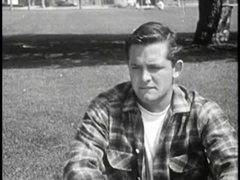 Young man in plaid shirt sitting outdoors, looking down.