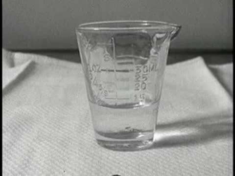 A measuring glass containing liquid on a white cloth on a table, vintage educational film.